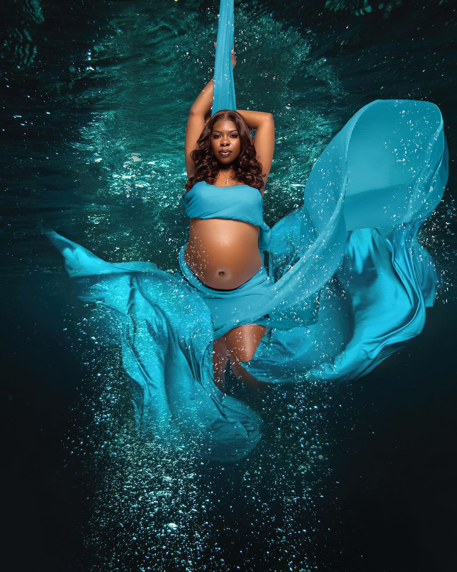 Maternity portrait in turquoise fabric with water splash effects against dark background.