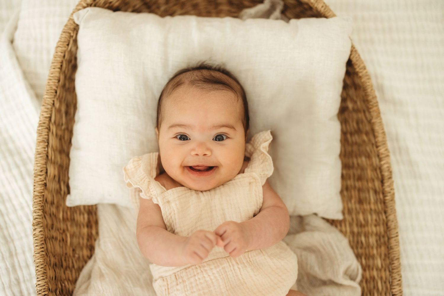 Happy baby wearing light pink outfit lies on white pillow in woven basket.