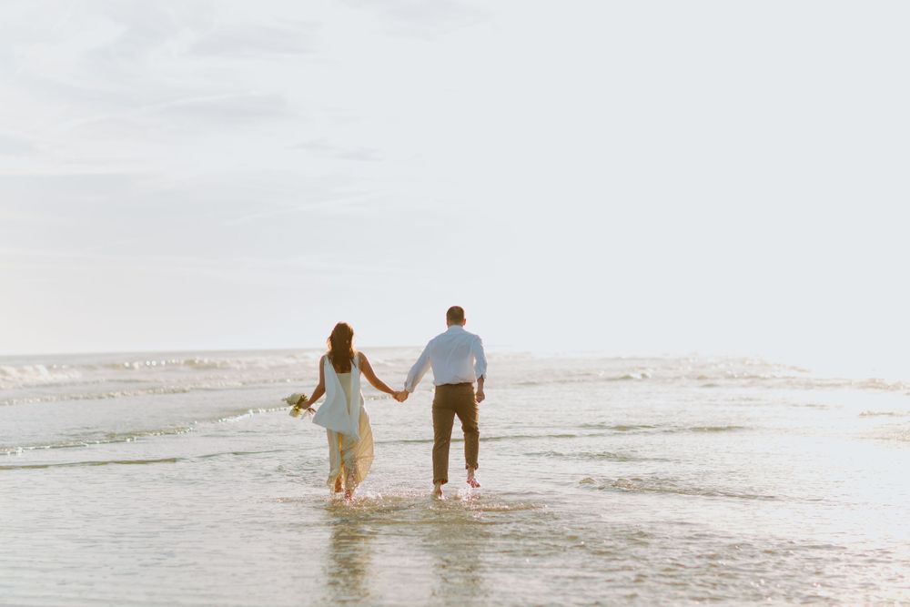 Couple holding hands while walking through shallow ocean water at sunset.