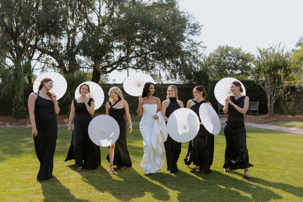 Bride in white gown with bridesmaids in black dresses holding white parasols on green lawn outdoors.