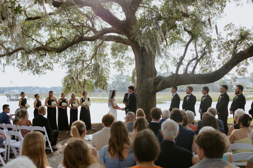 Outdoor wedding ceremony under a large oak tree by a waterfront, with bridal party in black and guests seated on white chairs.
