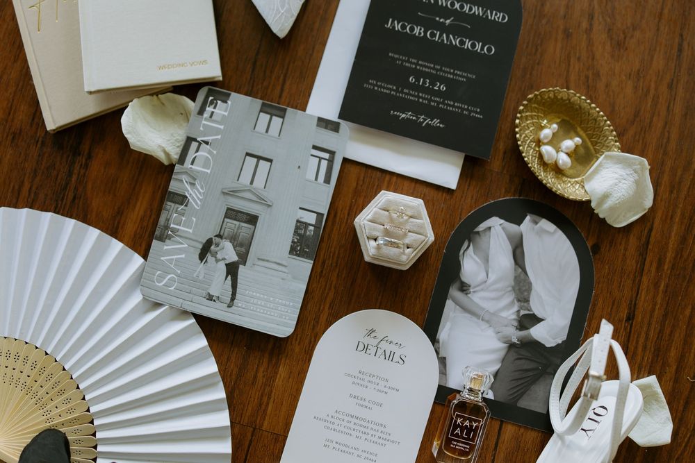 Elegant wedding stationery flatlay featuring black and white invitations, fan, perfume, and gold dish on wooden table.