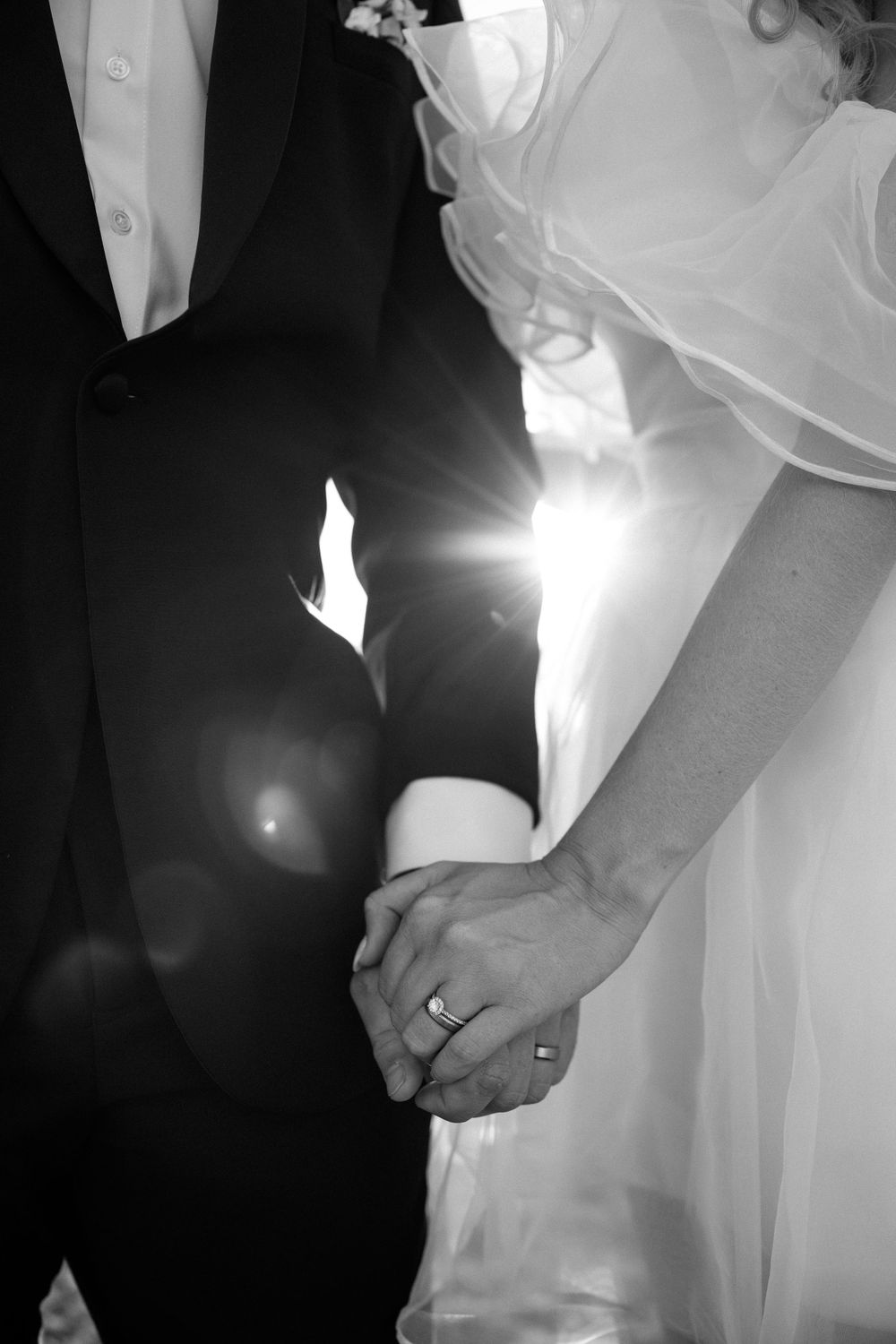 Black and white close-up of newlyweds holding hands with sunlight streaming between them.