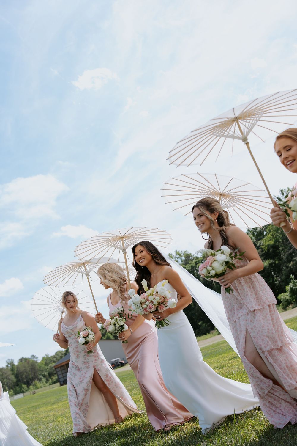 A wedding party poses playfully with white parasols on a grassy hill during a summer afternoon celebration.