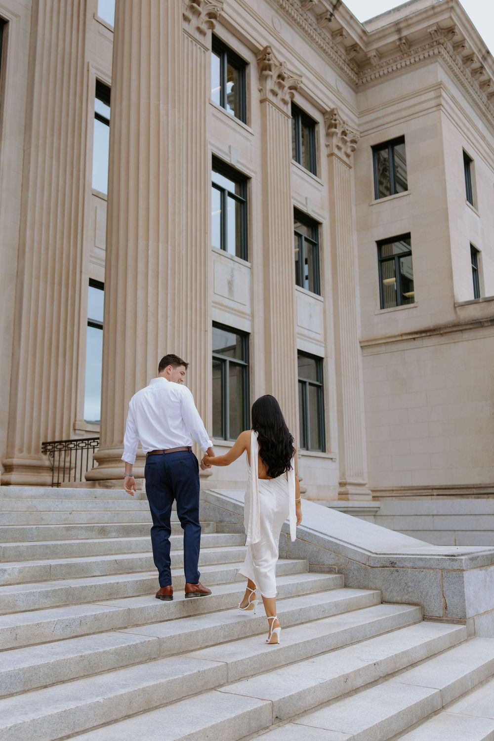 A couple walks up grand marble steps of a neoclassical building with tall columns and ornate architectural details.