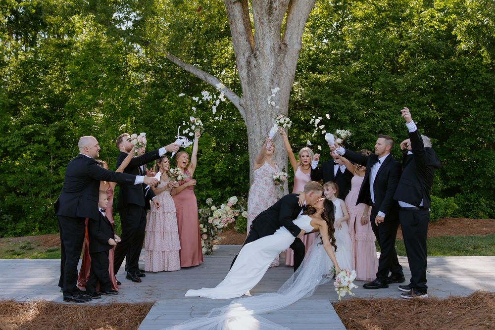 Wedding party celebrates with joyful poses during outdoor ceremony beneath large tree with blowing veil.
