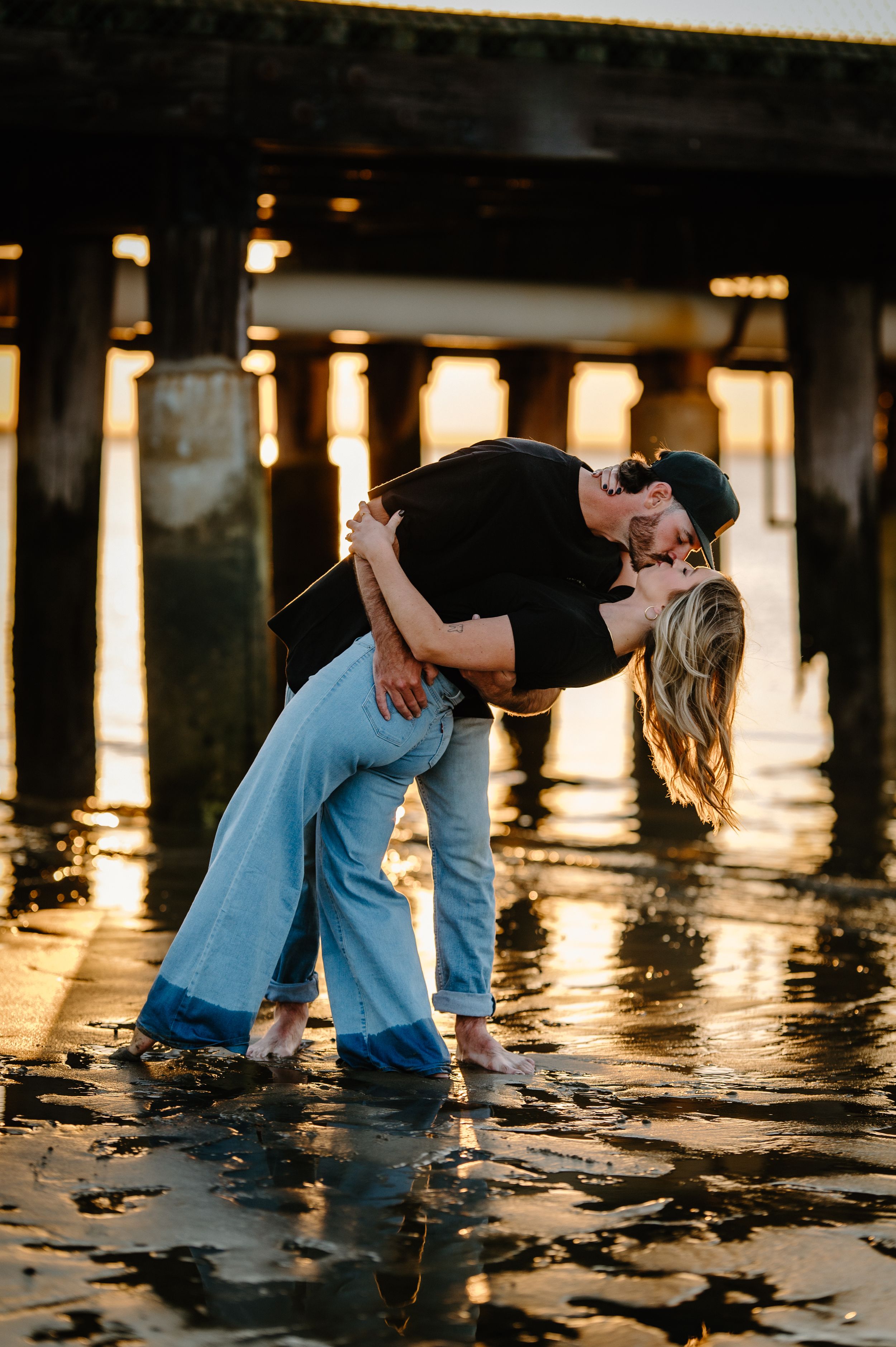 A romantic couple embraces by the water at sunset with reflections of light shimmering on the surface.