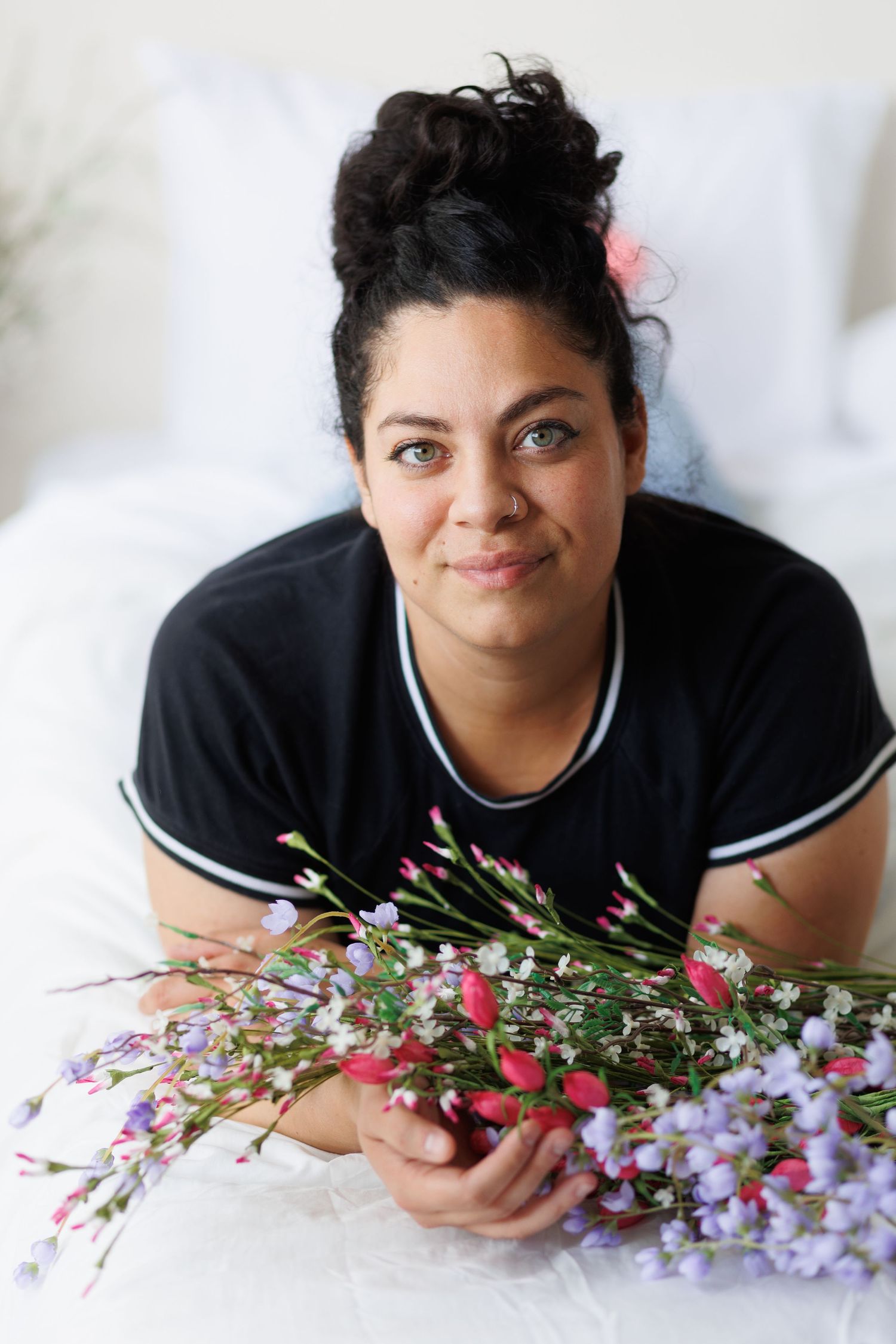 Woman with updo hairstyle lies on white bed holding colorful pink and purple flowers.