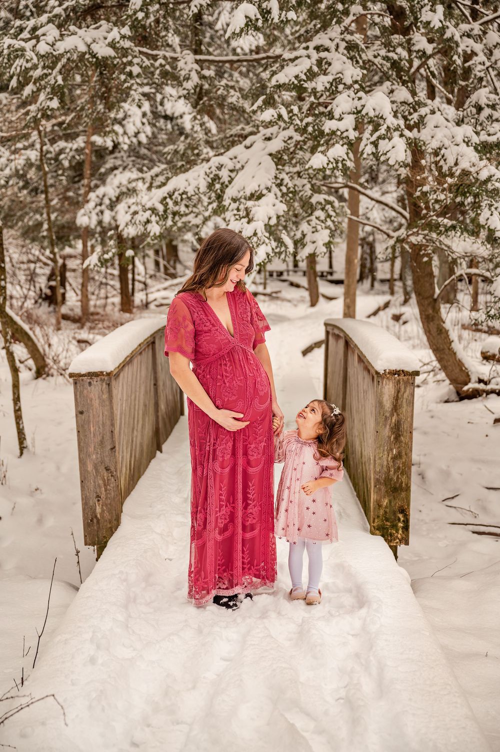 A pregnant woman in a red dress stands with a small child on a snow-covered wooden bridge in a winter forest setting.