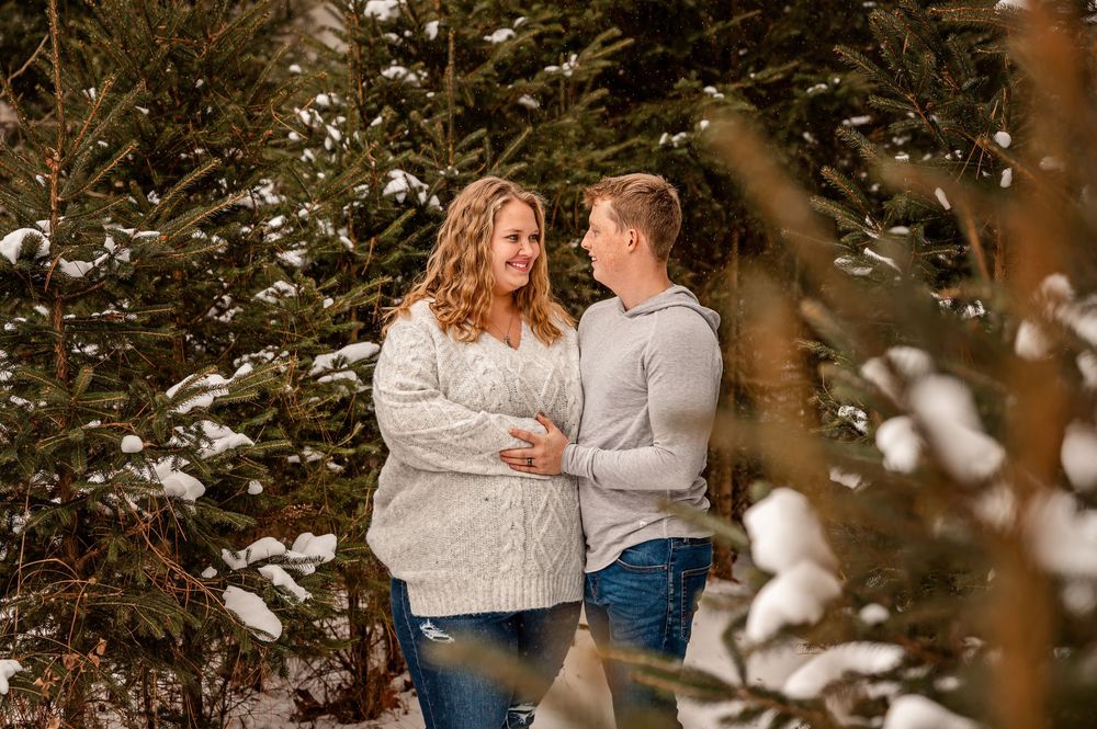 A couple in matching light sweaters share an intimate moment among snow-covered evergreen trees in a winter forest setting.