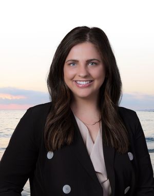 Professional headshot of a person in a black blazer with silver buttons smiling against an ocean sunset backdrop.