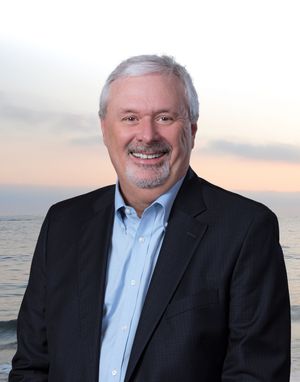 Professional headshot portrait of a person in a dark suit jacket and blue shirt against an ocean sunset background.