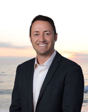 Professional business portrait of a person in a dark suit jacket and white shirt against an oceanfront sunset backdrop.