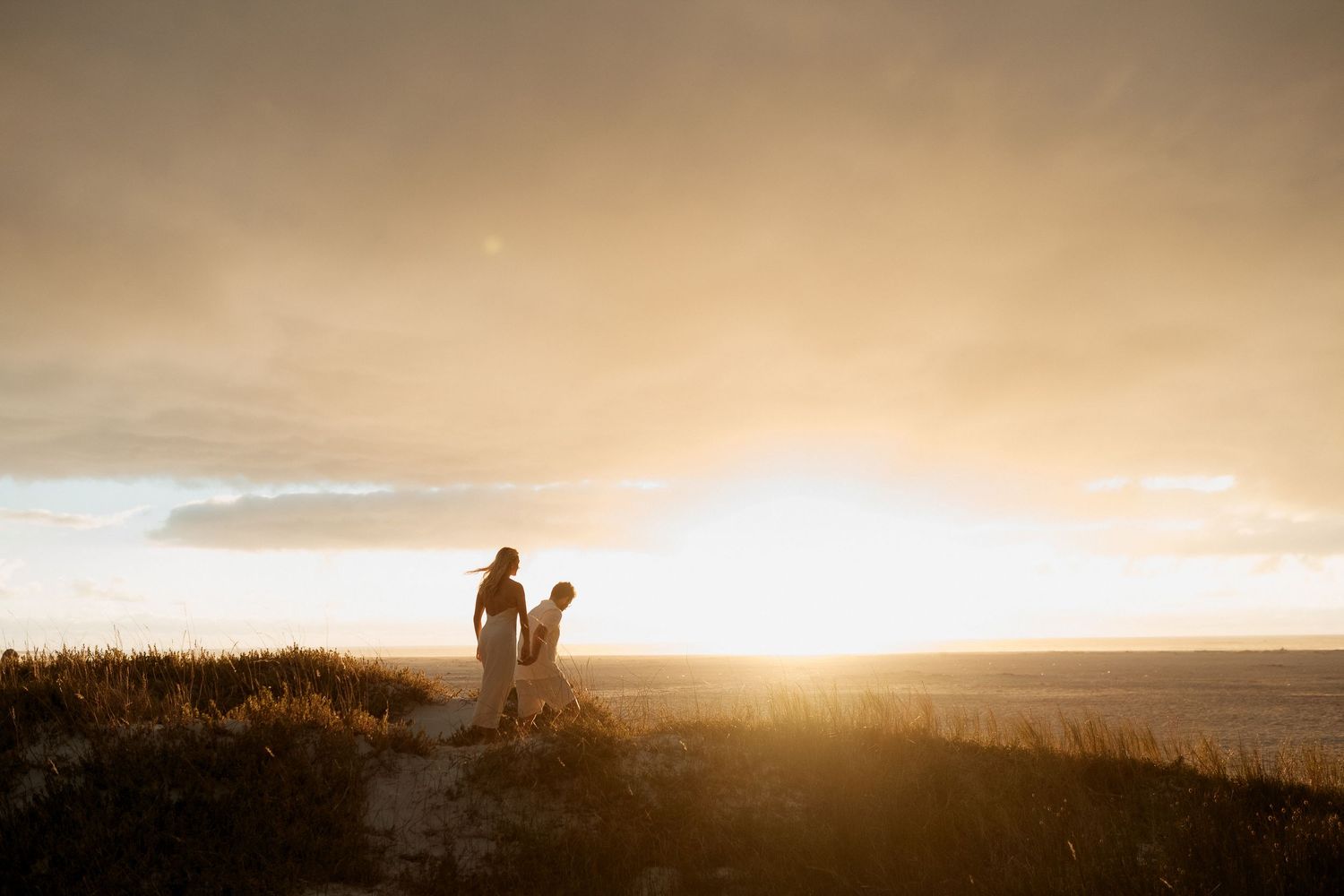 Two silhouettes stand on a sandy dune at golden sunset, bathed in warm glowing light against a dramatic sky.