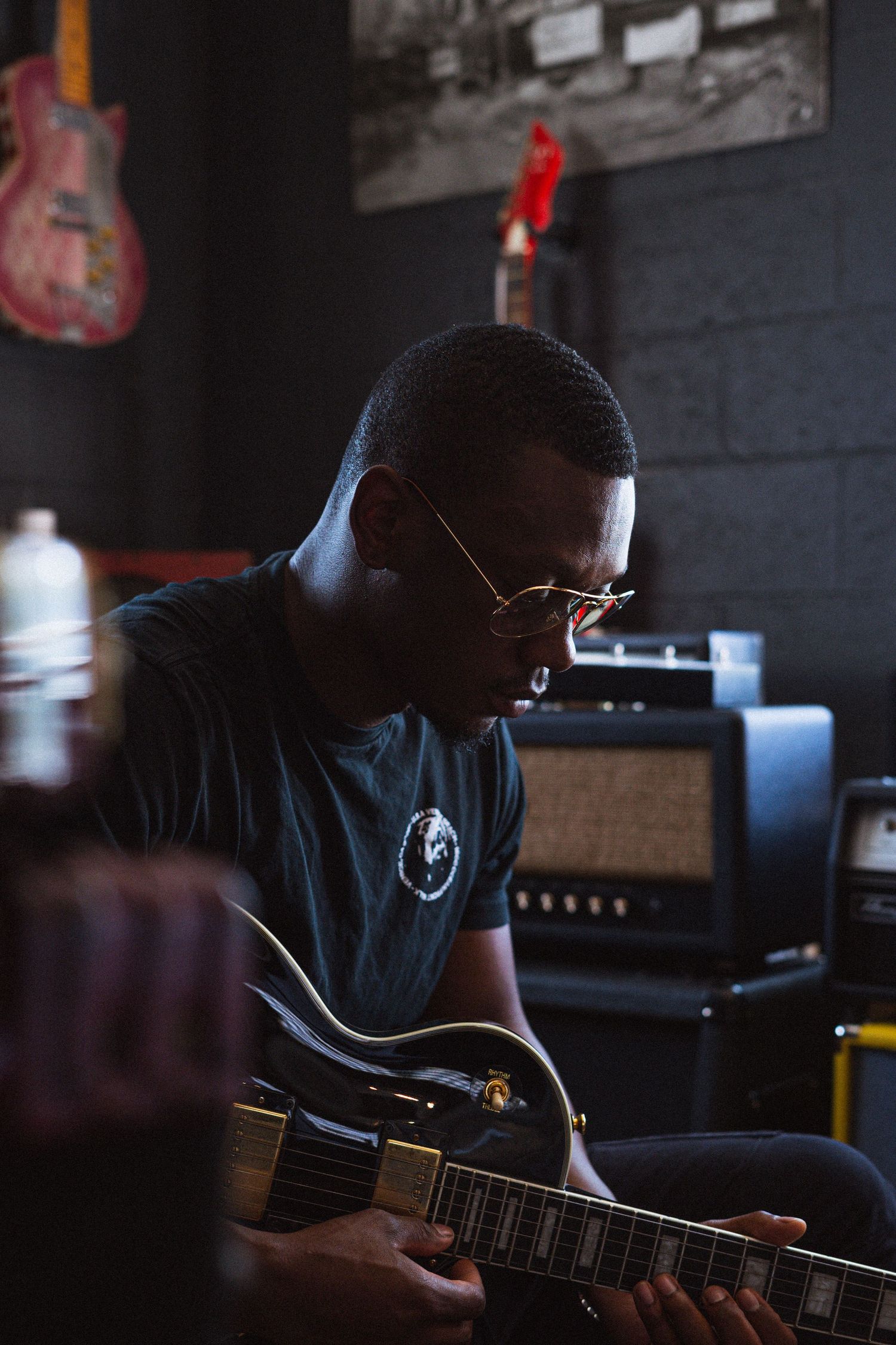 A musician plays guitar in a dimly lit studio space with brick walls.