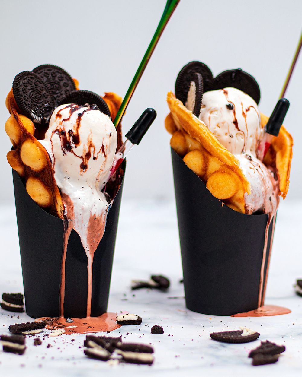 Two black cups filled with melting ice cream scoops, Oreo cookies, waffle pieces, and chocolate drizzle on white surface.