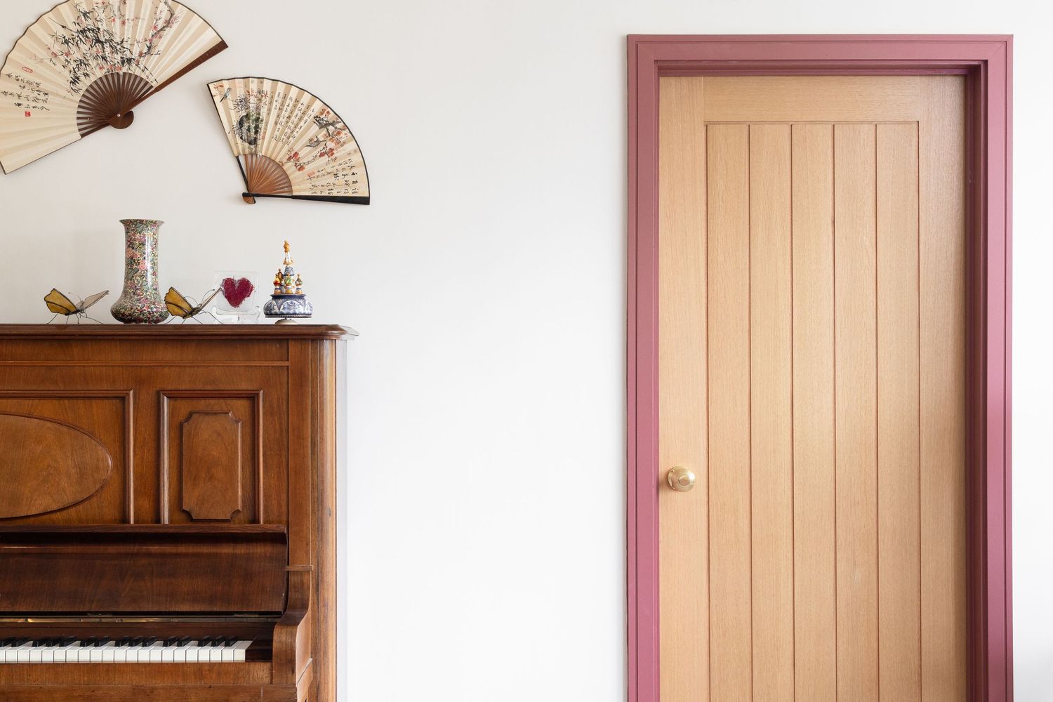 Hallway detail with upright piano, decorative wall fan and pink-framed wooden door, eclectic interior decor photography