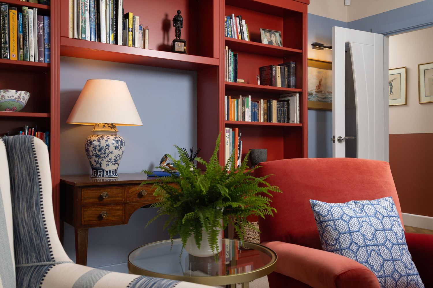 Cosy living room reading nook with red bookshelves, armchair, table lamp and fern plant, interior decor photography