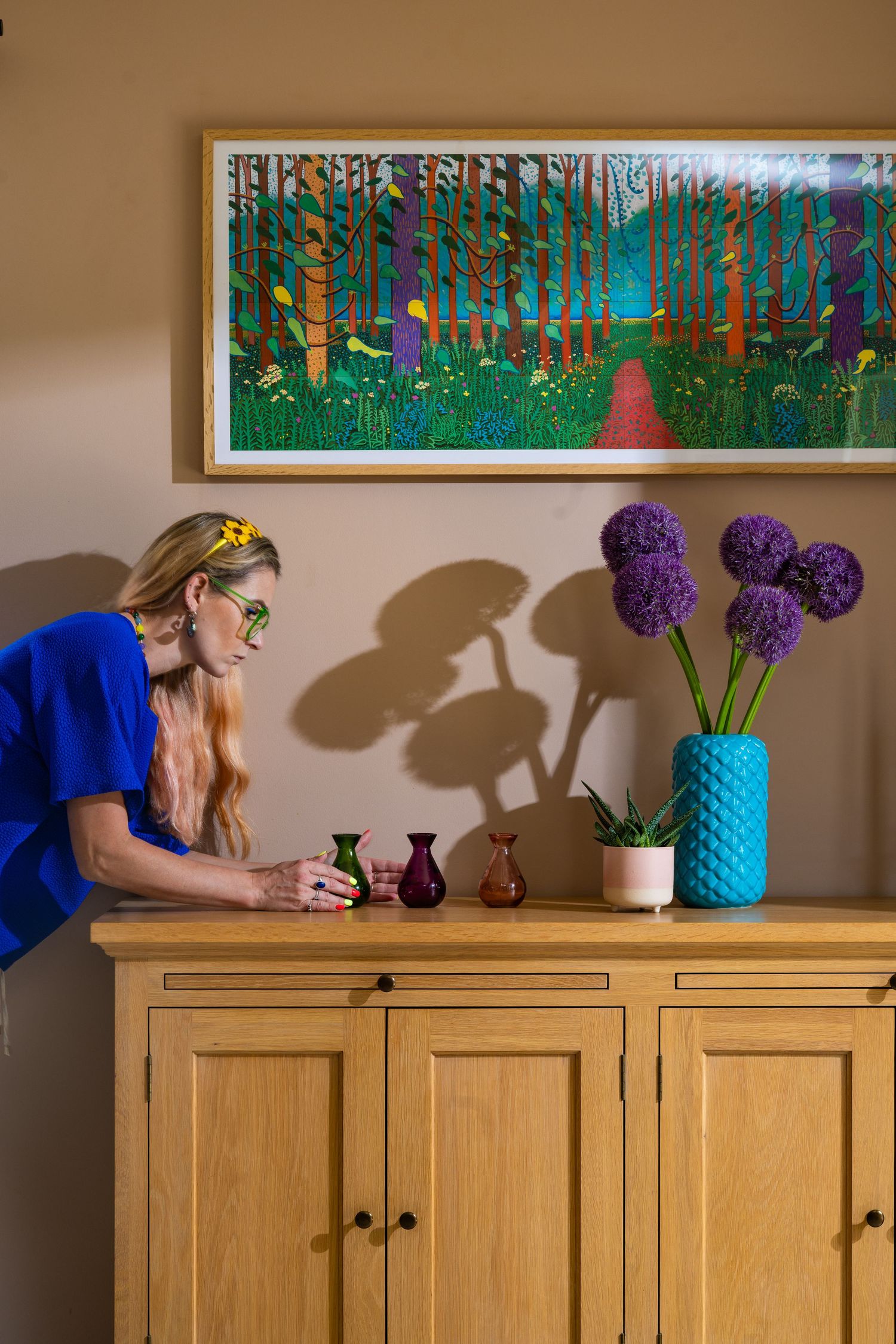 Wooden sideboard with colourful framed artwork, purple alliums in teal vase and decorative objects, interior styling photography