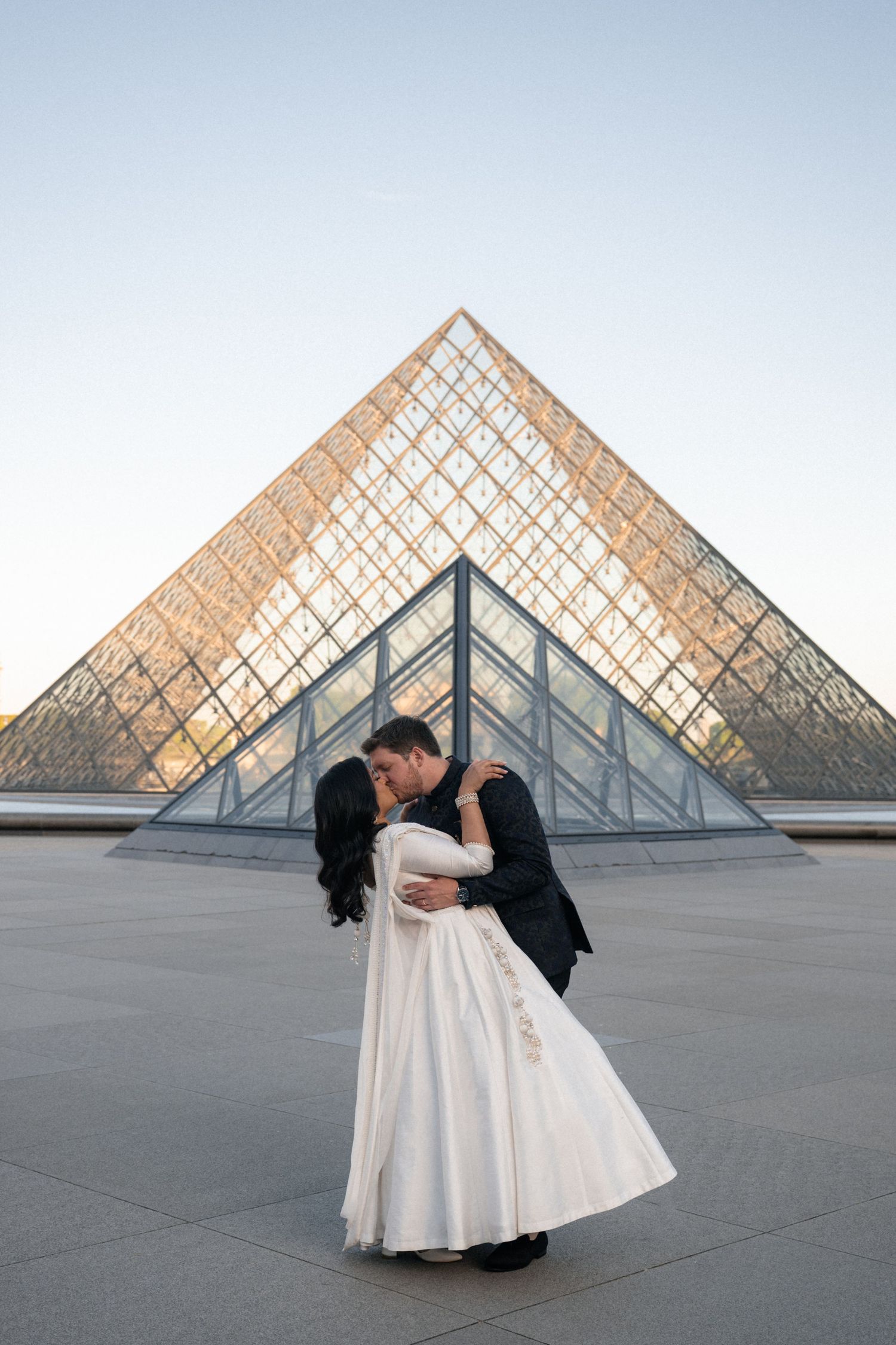A stylish couple poses together in formal attire under classical architecture.