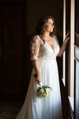 Bride in flowing white lace dress poses dramatically by window light holding bouquet.