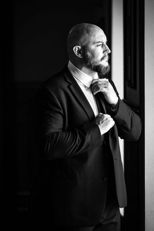 Groom in black tuxedo stands thoughtfully near window in dramatic lighting.