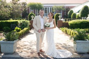 A couple in formal attire dances together on a sunlit garden pathway surrounded by manicured hedges.