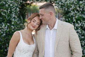 A romantic couple embraces in front of a lush green hedge wall during an outdoor photoshoot.
