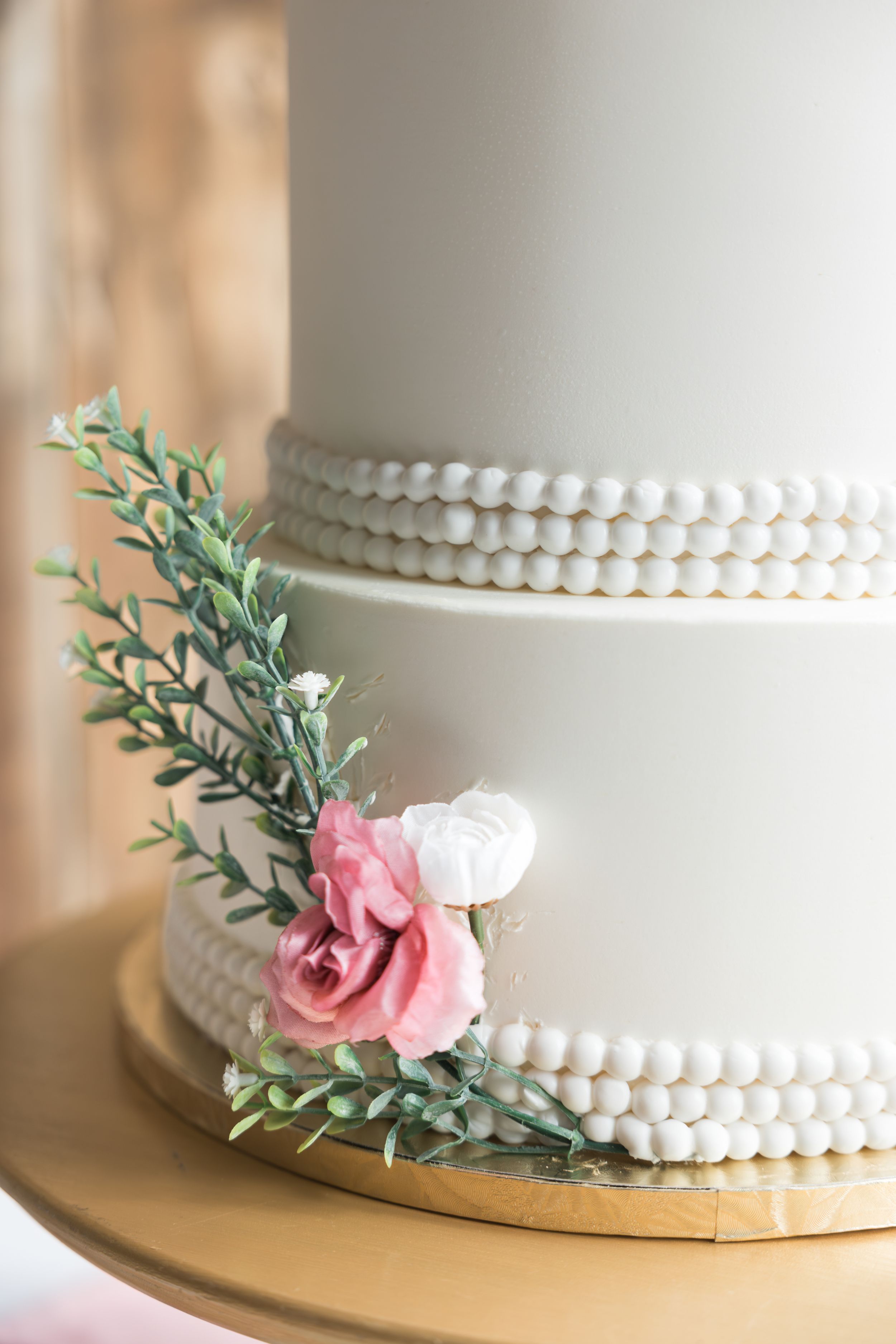 White wedding cake with delicate pearl border and pink rose accent on gold cake stand.