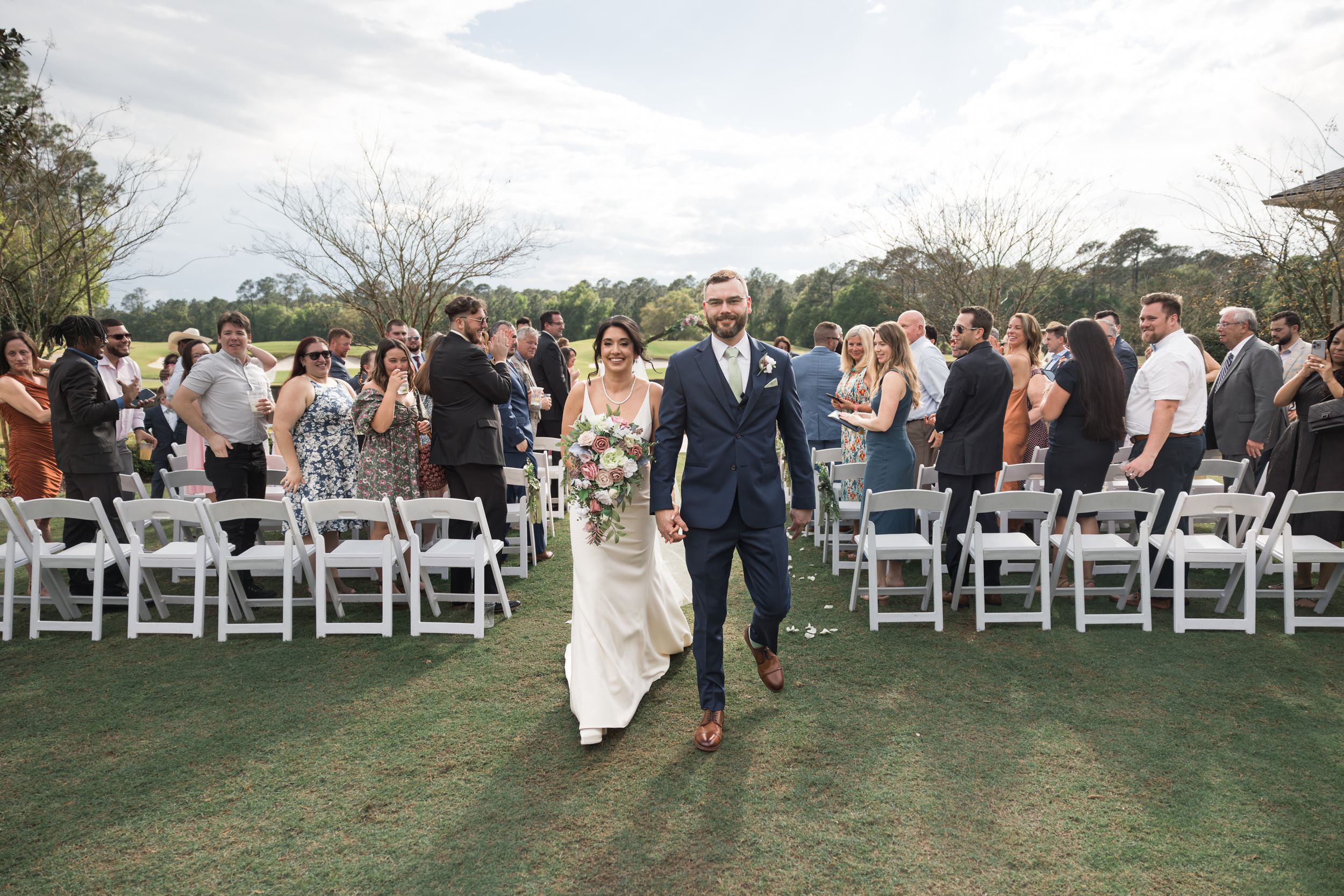 Wedding guests seated in white chairs on a lawn during an outdoor ceremony.