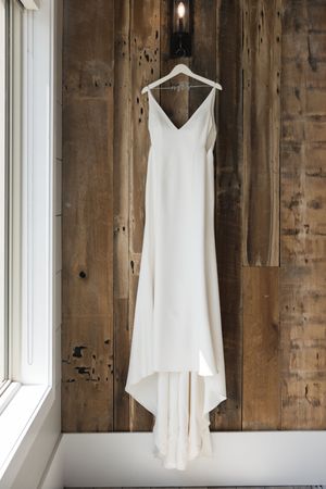 A white wedding dress hangs against a rustic wooden wall with natural window lighting.