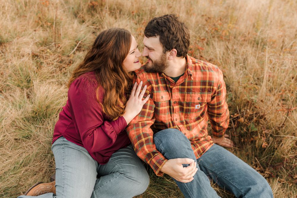 A couple in casual fall attire sits together in a golden grass field during sunset.