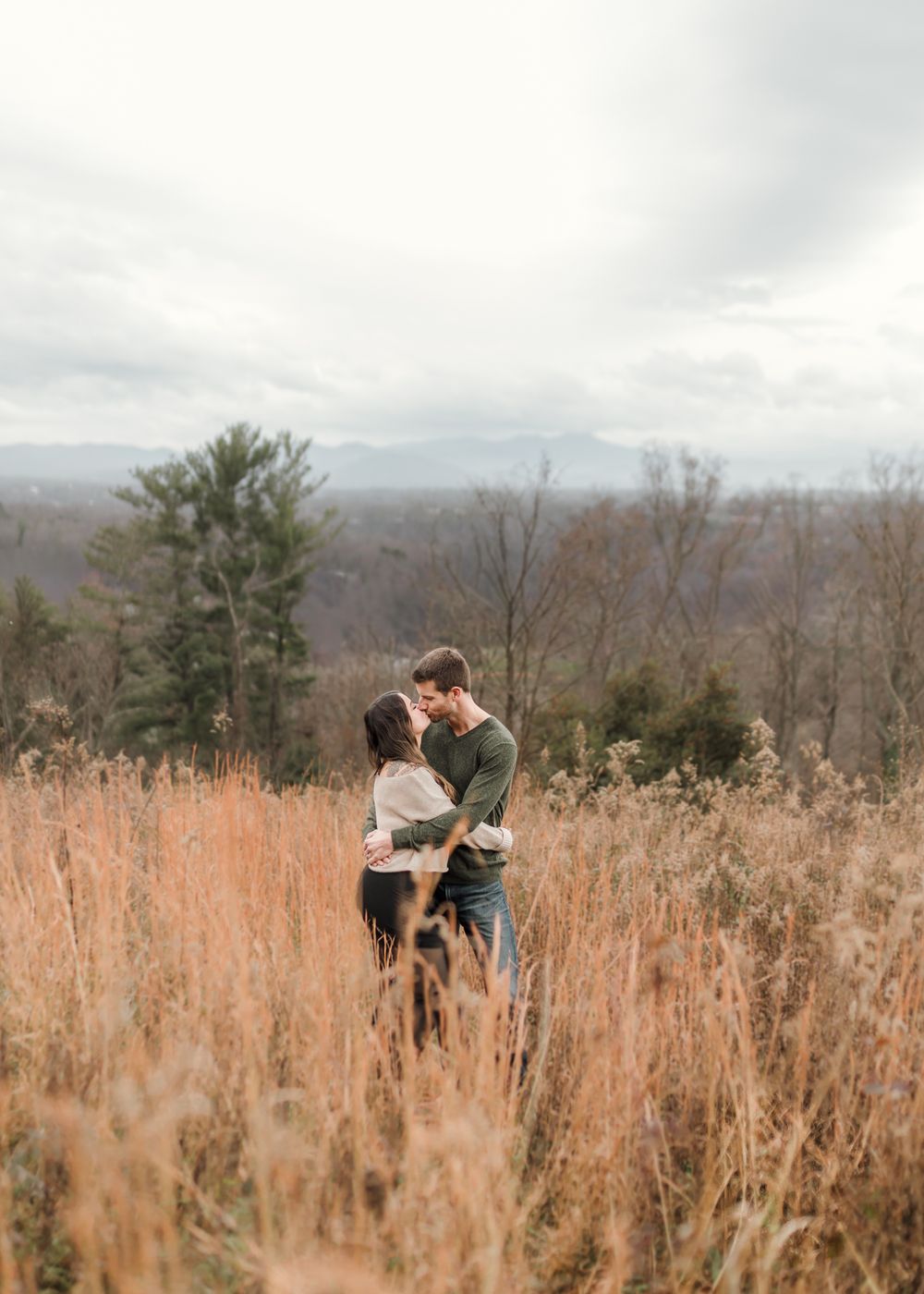 A couple shares an intimate moment among tall autumn grasses in a scenic mountain overlook setting.