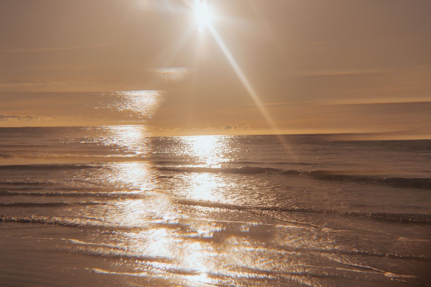 Golden sunset reflecting off shallow ocean waves on a beach with bright sun flare.