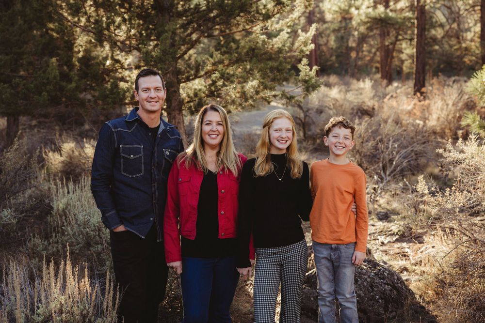 A family stand together outdoors in casual attire against a backdrop of autumn foliage and brush.