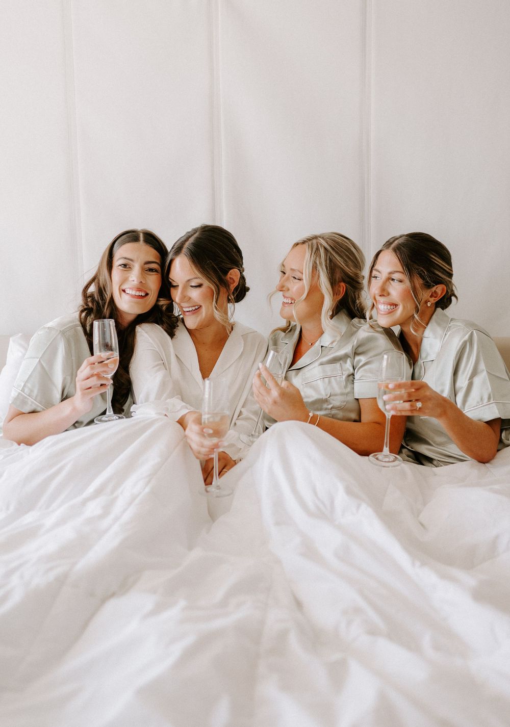 Friends in white robes share champagne and laughter while relaxing in bed.