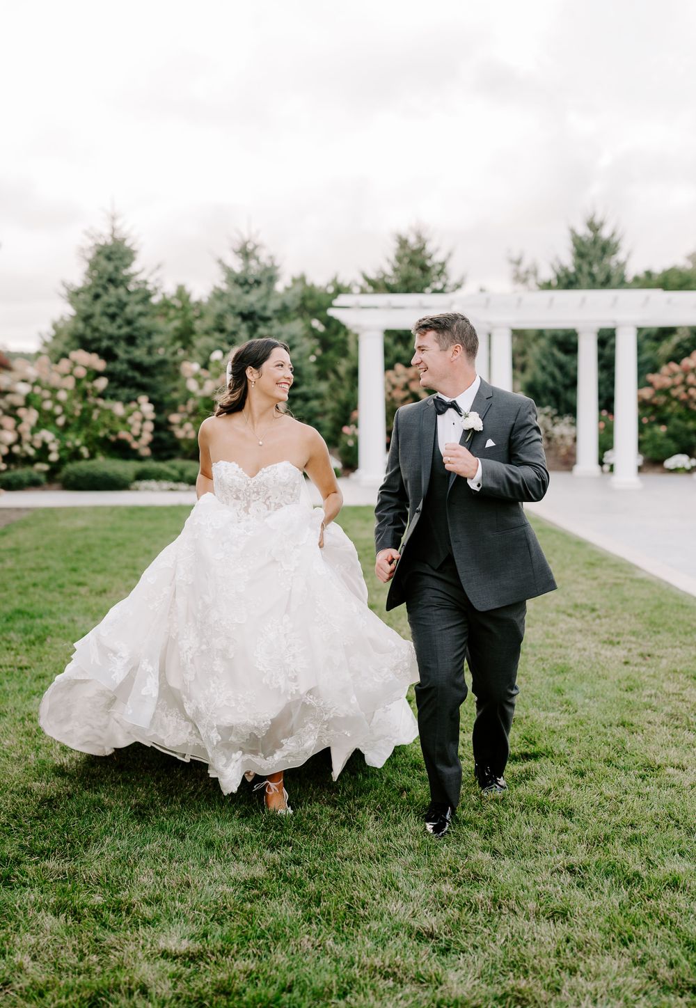 A couple runs joyfully across a lawn with a white pergola in the background during their wedding celebration.