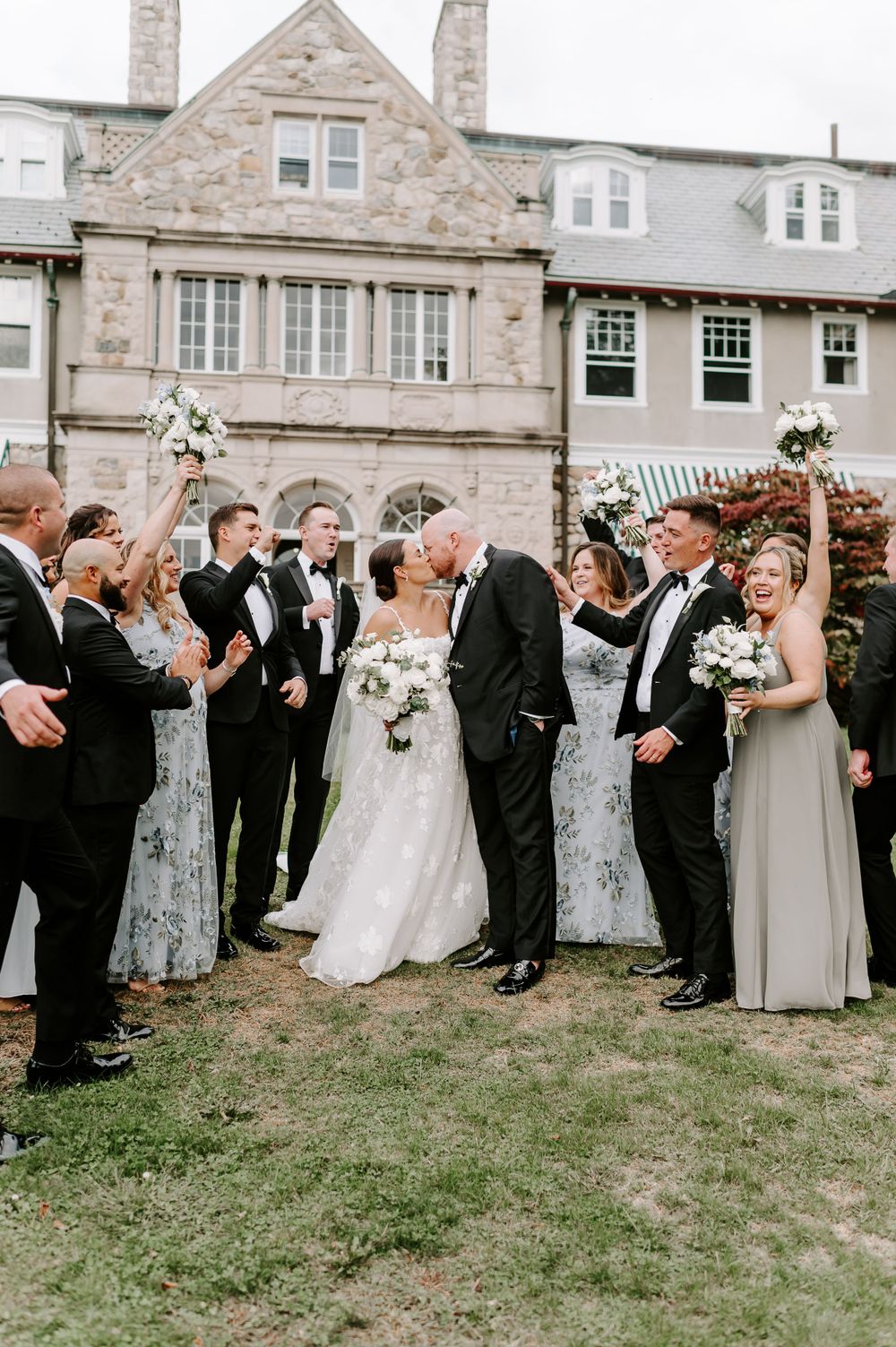Large wedding party celebrating with joyful poses outside a stone building.