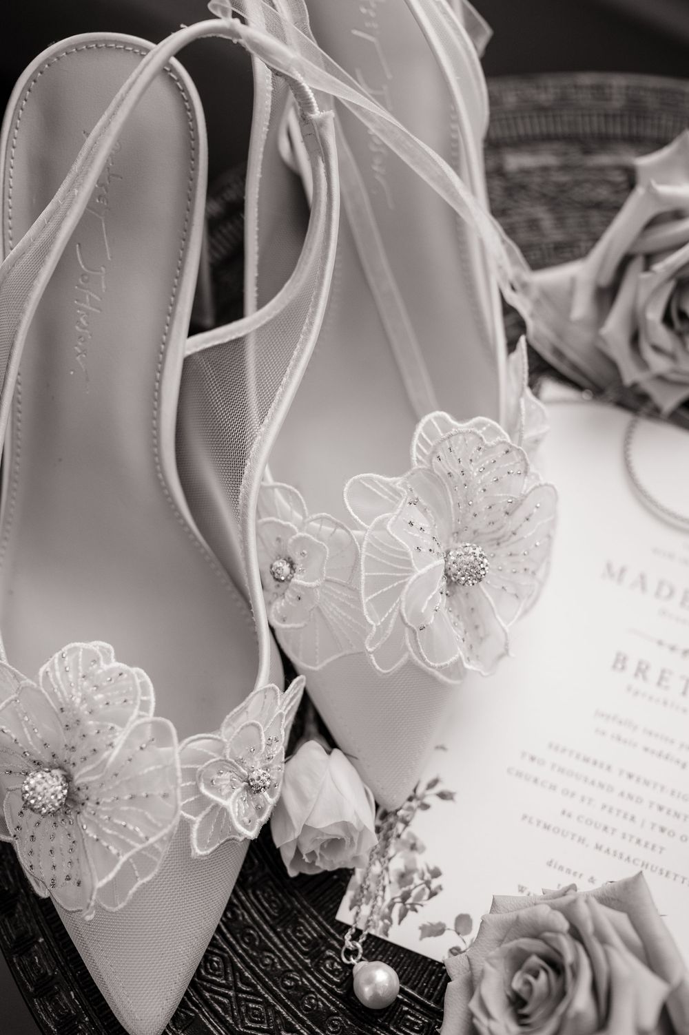 Elegant white bridal shoes with floral appliques displayed next to wedding invitations and roses.