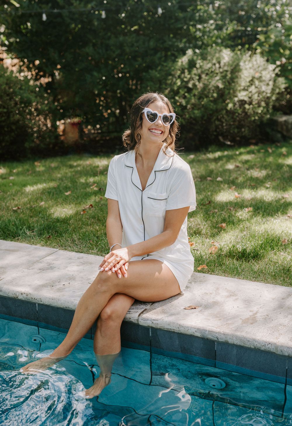 A person in white pajamas and sunglasses sits poolside with feet in the water.