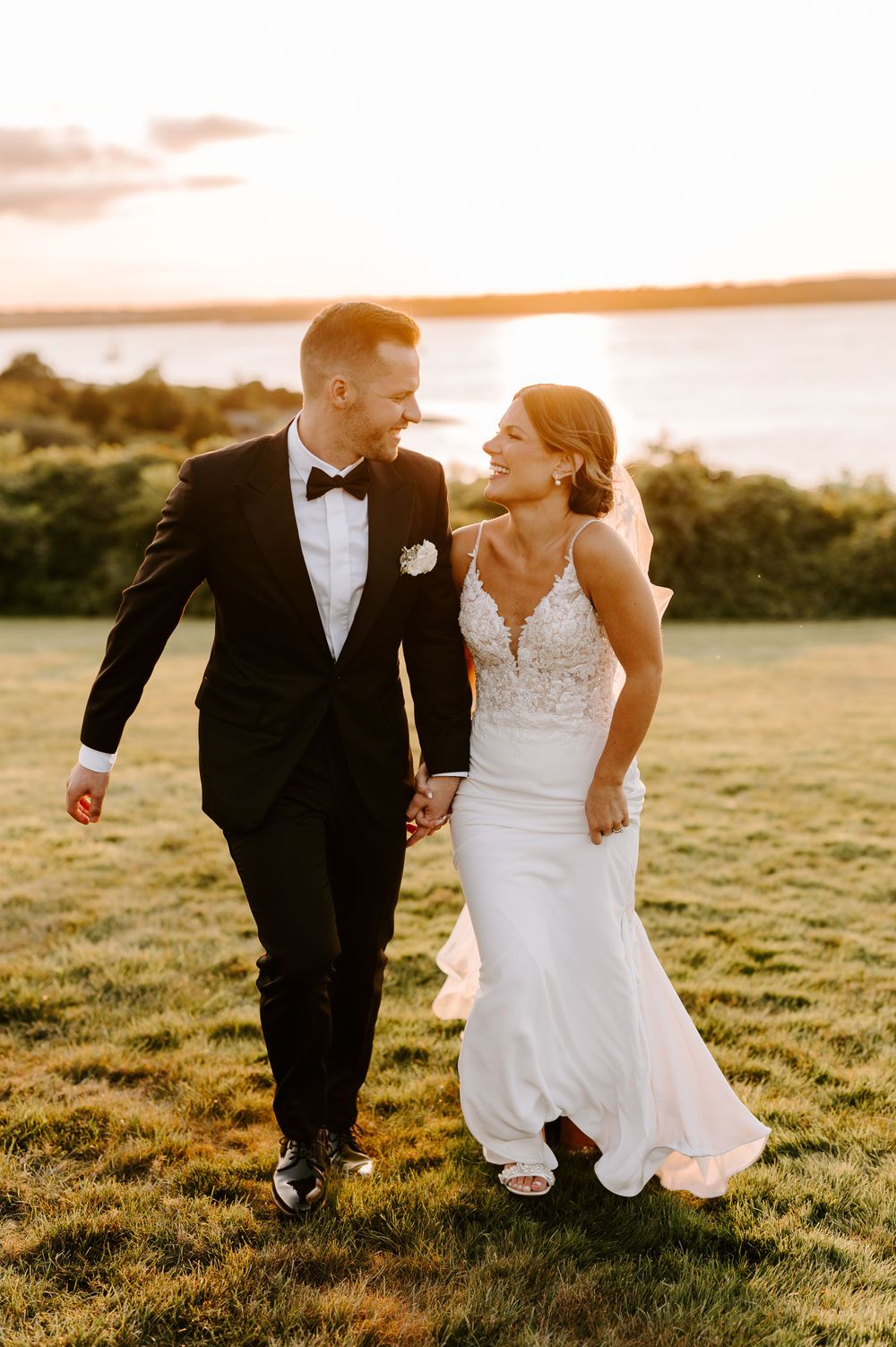 Romantic couple in wedding attire walking and laughing together in a sunlit field at golden hour.