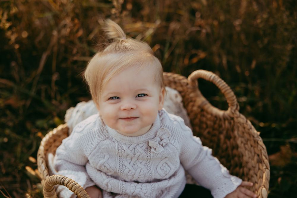 Small baby wearing white knit sweater sitting in woven basket outdoors with autumn background.