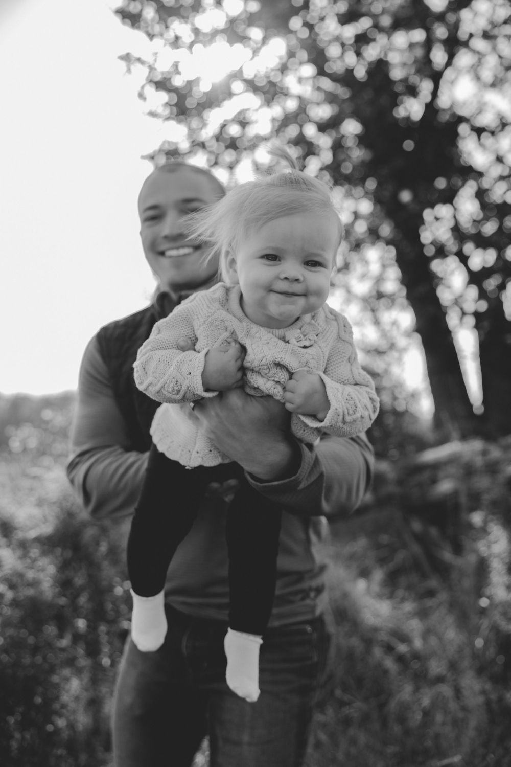 Black and white portrait of an adult holding a smiling baby outdoors with bokeh lights in the background.