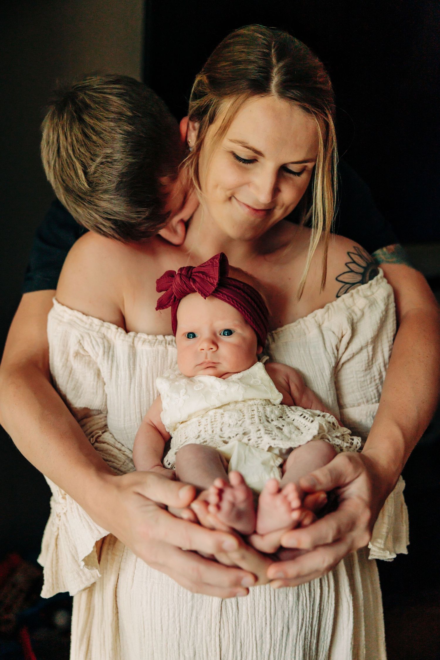 Tender family moment captured in soft light with baby in white lace dress.