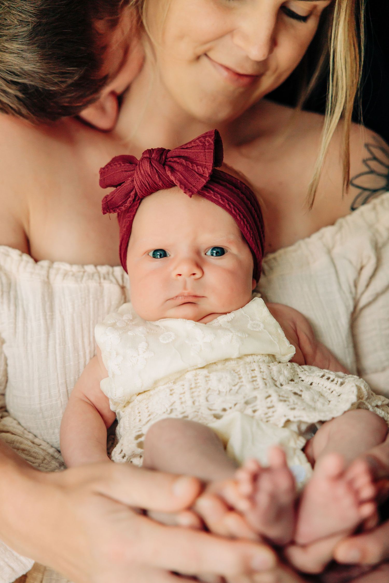 Infant wearing white lace dress and burgundy bow headband rests against soft fabric.