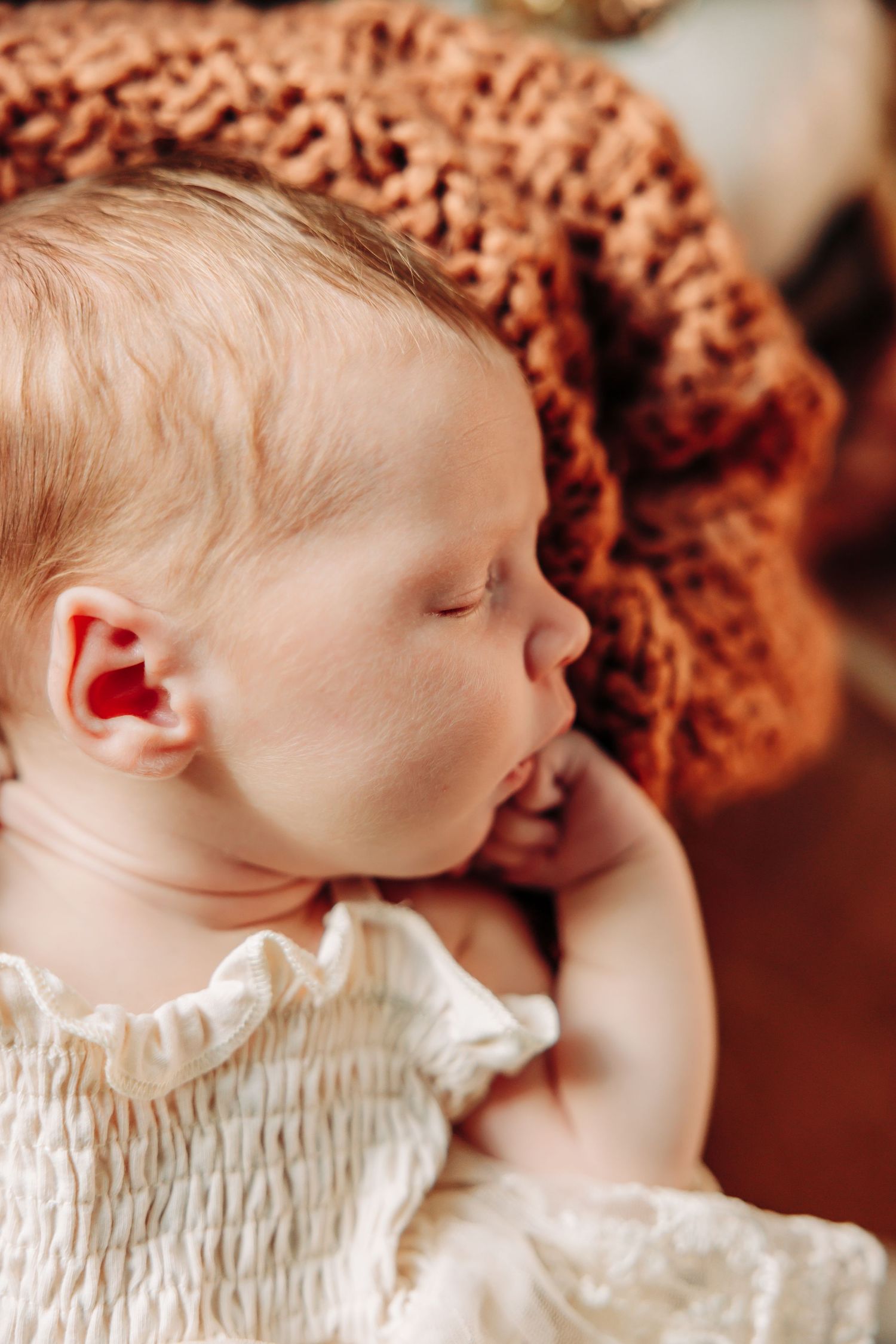 Newborn baby napping on soft peach-colored knit blanket in white dress.