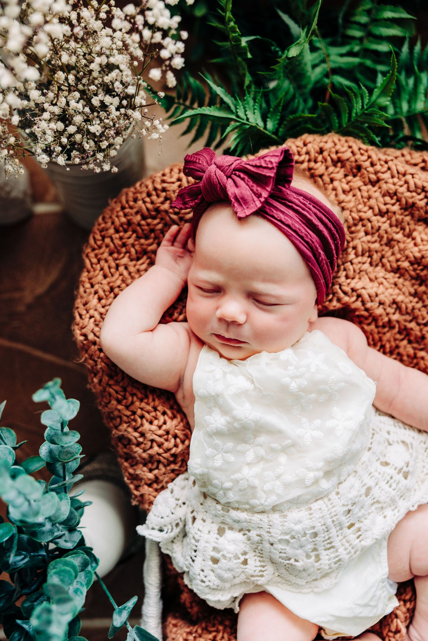 Sleeping baby in white lace dress and burgundy headband on knit blanket with florals.