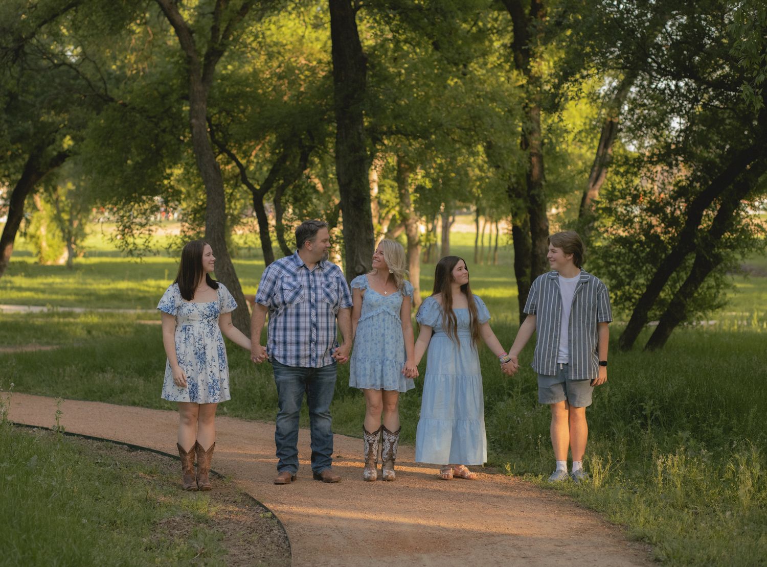 Family of five holding hands walking along a sunlit wooded path in golden afternoon light.