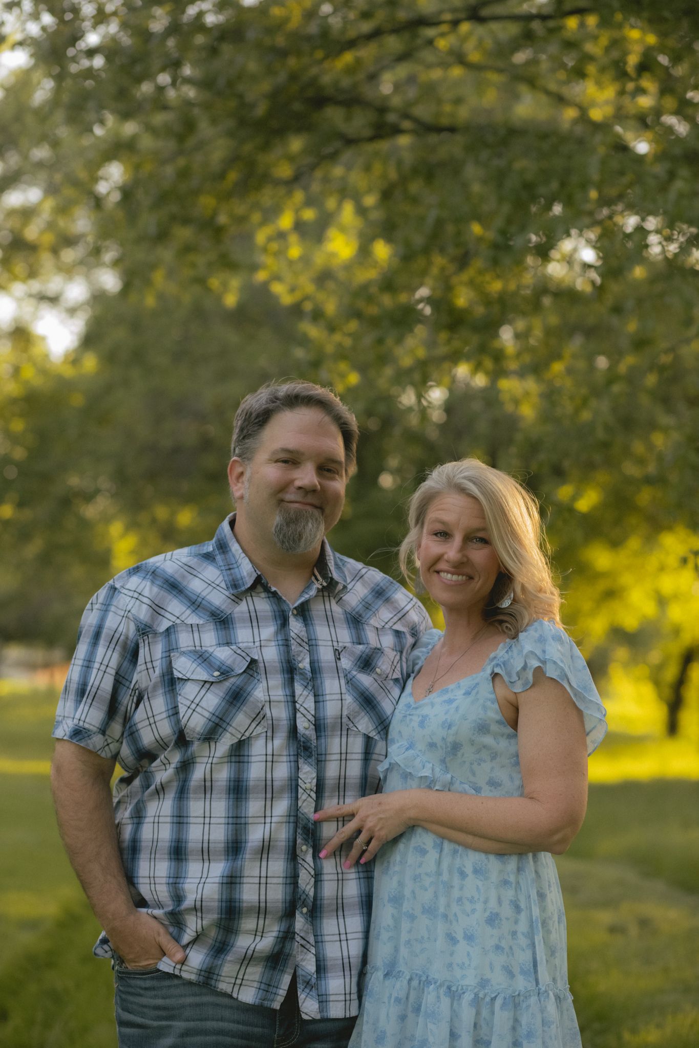 Couple smiling outdoors in golden hour light, man in plaid shirt and woman in blue floral dress.