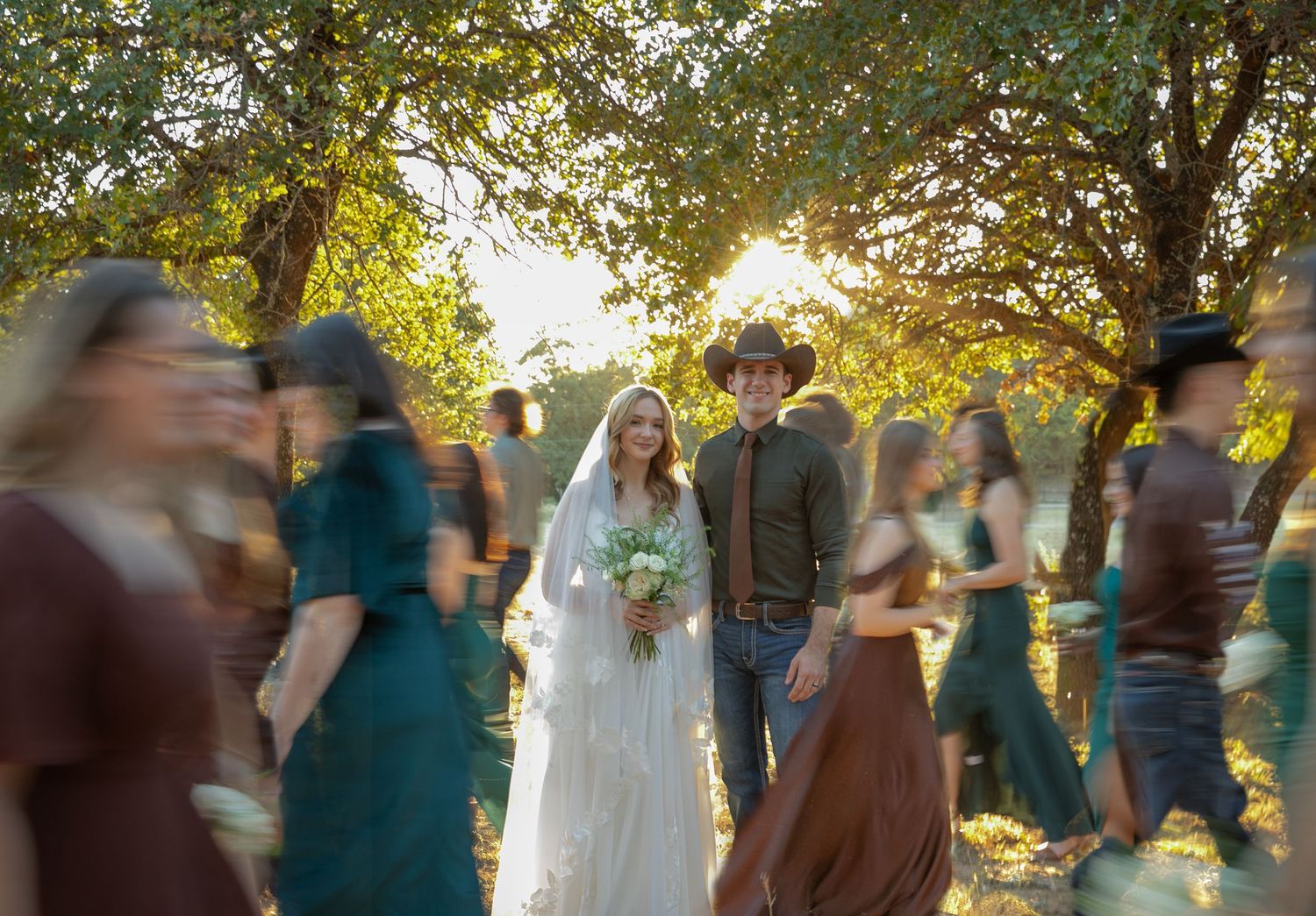 A bride and groom walk through a crowd of guests in a sunlit outdoor wedding surrounded by lush green trees.